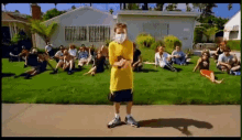 a young boy wearing a face mask is standing in front of a group of people sitting on the grass .