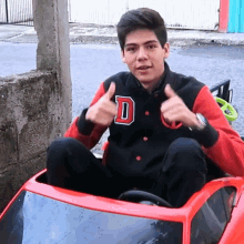 a young man is giving a thumbs up while sitting in a red toy car with the letter d on his jacket