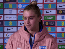 a woman wearing a pink jacket stands in front of a wall of advertisements including google cloud and m & s