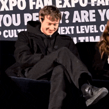 a man sits on a couch with his legs crossed in front of a sign that says ' expo fan expo ' on it