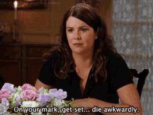 a woman is sitting at a table with flowers and saying on your mark get set die awkwardly