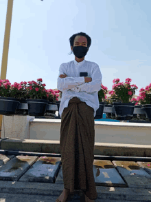 a man wearing a mask stands with his arms crossed in front of flowers