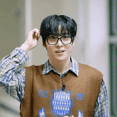 a young man wearing glasses , a vest and a plaid shirt is adjusting his hair .
