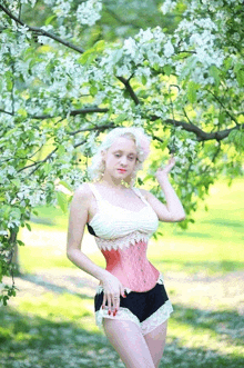 a woman in a corset and shorts stands under a tree with white flowers