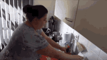 a woman in a blue shirt is washing dishes in a kitchen with a carton of aquafina on the counter