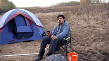 a man sits in a chair in front of a tent with a bucket next to him that says fire