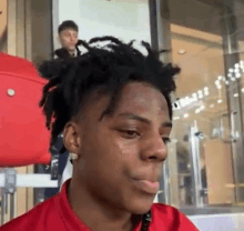 a young man with dreadlocks and a red shirt is sitting in front of a glass door .