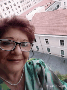 a woman wearing glasses and a pearl necklace is smiling in front of a building with the date 2014/12/2 on the bottom