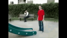 a man in a red shirt is standing in front of a slide in a park while a woman sits on a bench .