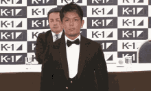 a man in a tuxedo stands in front of a wall that says k- 1