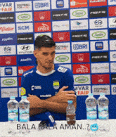 a man in a blue shirt sitting at a table with bottles of water in front of a wall with logos for companies such as bri