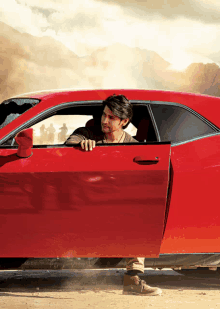 a man leaning out of a red car with mountains in the background