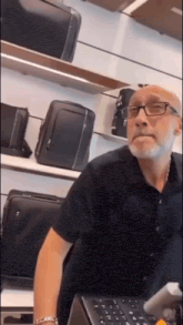 a man with glasses and a beard is sitting at a counter in front of a wall of luggage