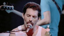 a man with a mustache singing into a microphone with another man playing a guitar in the background