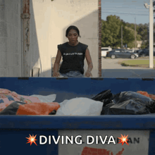 a woman in a diving diva shirt stands next to a dumpster