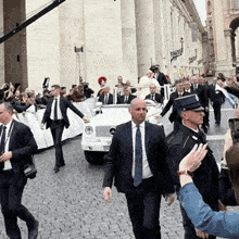 a man in a suit and tie is walking in front of a white car with a license plate that starts with s on it