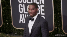 a man in a tuxedo and bow tie smiles on a red carpet in front of a sign that says golden globe awards