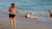 a woman in a swimsuit is standing on a beach next to a man and a woman laying in the water .