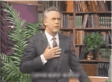 a man in a suit and tie is standing in front of a bookshelf with his hand on his chest .