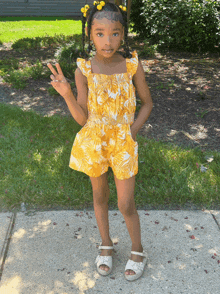 a little girl wearing a yellow jumpsuit and white sandals is giving a peace sign