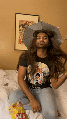 a man wearing a queen shirt sits on a bed next to a bag of lays