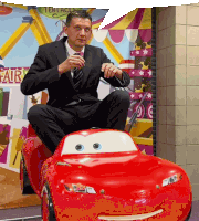 a man in a suit and tie is sitting on a toy car with the number 99 on the front