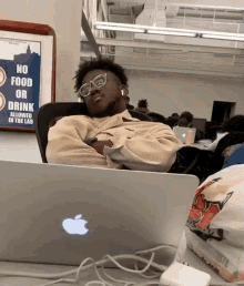 a man sleeping in front of an apple laptop with a sign behind him that says no food or drink allowed in the lab