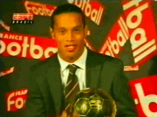 a man in a suit and tie is holding a soccer ball in front of a wall that says " football "