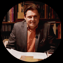 a man in a suit sits in front of a bookshelf with a book called art of war on it