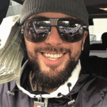 a man with a beard wearing sunglasses and a beanie is smiling while holding a stack of money .