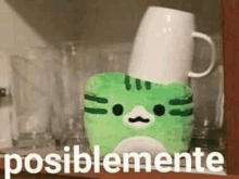 a green cat stuffed animal is sitting on top of a white coffee mug .