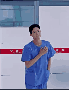 a man in a blue scrub is standing in front of a hospital door with his hand on his chest .