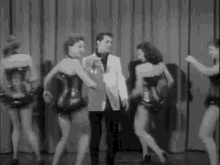 a man in a suit is standing in front of a group of women dancing on a stage .