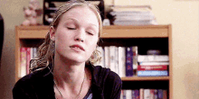 a woman is sitting in front of a bookshelf with her eyes closed and a necklace around her neck .