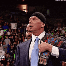 a man in a suit and tie holds a wrestling belt over his shoulder