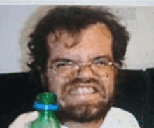 a man with a beard and glasses is holding a green bottle and making a funny face .