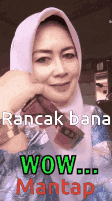 a woman wearing a hijab is holding a cell phone and says wow mantap on the bottom