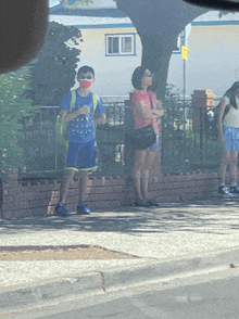 a boy wearing a mask stands on a sidewalk waiting for a bus