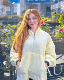 a woman with long red hair is wearing a yellow sweatshirt and standing in front of a sign .