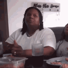 a woman is giving a thumbs up while sitting at a table in front of a sign that says bless this home