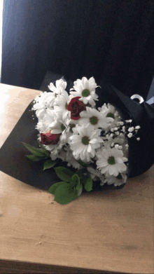 a bouquet of white daisies and red roses wrapped in black paper on a table