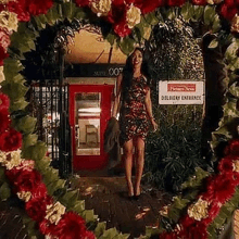 a woman in a red dress is standing in front of a red phone booth surrounded by flowers .