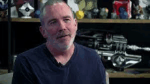 a man with a beard and a blue shirt is sitting in front of a shelf .