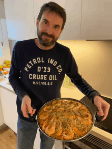 a man wearing a petrol ind co crude oil sweatshirt holds a pan of food