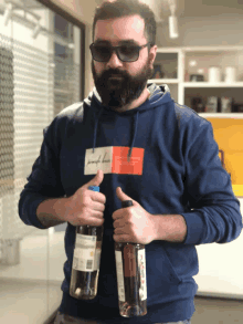 a man with a beard wearing sunglasses and a blue hoodie holds two bottles of alcohol