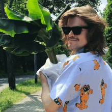 a man wearing sunglasses and a garfield shirt holds a banana plant