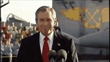 a man in a suit and tie is standing in front of two microphones in front of a plane .