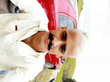 a bald man with a mustache and a white shirt is smiling in front of a red car