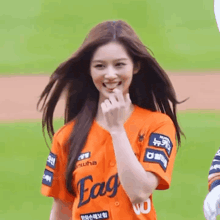 a woman in an orange baseball jersey is smiling and making a heart shape with her finger .