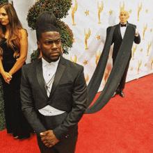 a man in a tuxedo stands on a red carpet with a woman behind him